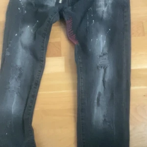 Svarta jeansbyxor från Dsquared2 med slitningar - Säljer ett par svarta jeansbyxor från Dsquared2 med slitningar, färgstänk och dragkedjedetalj. Byxorna har en smal passform och unika detaljer vid fickorna. Perfekta för dig som gillar en edgy stil.