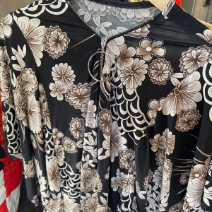 Omlottklänning med dragkedja - Snygg svart klänning med stora beige och vita blommor, omlottmodell och dragkedja framtill med rund metallring. Klänningen har korta ärmar och ett flowy fall, perfekt för dig som gillar statement-mönster.