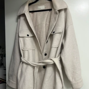 Beige overshirt med bälte från NA-KD - Snygg beige overshirt från NA-KD med bälte i midjan och stora bröstfickor. Jackan har knappar framtill och på ärmsluten samt klassisk krage. Perfekt för lager-på-lager och enkel att styla till olika outfits.