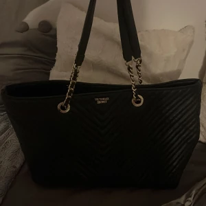 Svart quiltad axelväska från Victoria's Secret - Snygg svart axelväska från Victoria's Secret med quiltad design och kedjedetaljer på axelbanden. Perfekt storlek för att få plats med allt du behöver. Stängs upptill och har en lyxig känsla.