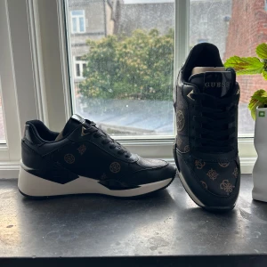 Svarta sneakers från Guess med mönster - Säljer ett par svarta sneakers från Guess med bruna logomönster och vita sulor. Skorna har snörning och en sportig design med rund tå. Perfekta för dig som vill ha en snygg och trendig vardagssko. Aldrig använda, ligger i originalförpackning med priset på kartongen.