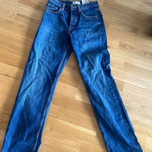 Blå raka jeans från Zara - Klassiska blå jeans från Zara med raka ben och normal passform. Jeansen har fem fickor, silverfärgade knappar och tydliga sömmar. Perfekta till vardags och lätta att matcha med olika stilar.