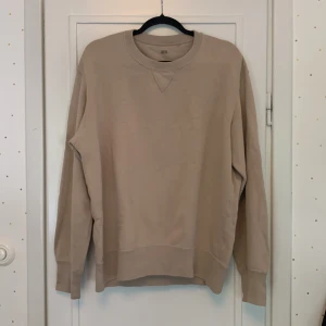Beige sweatshirt från Uniqlo - Säljer en stilren beige sweatshirt från Uniqlo storlek L. Tröjan har rund halsringning med klassisk V-detalj framtill och långa ärmar. Tillverkad i mjuk bomullsblandning med ribbade muddar vid ärmslut och nederkant. 