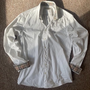 Vit burberry skjorta - Vit skjorta från Burberry med klassiska rutiga detaljer på insidan av kragen och ärmsluten. Skjortan har lång ärm och knäppning framtill. Perfekt för dig som gillar stilrena plagg med en twist. Skick: 7,5 bra skick, köpt secondhand. Priset är diskuterbart som alltid