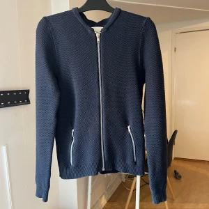 Mörkblå stickad kofta  - Säljer en mörkblå stickad jacka från Studio Total med silverfärgad dragkedja framtill och vid fickorna. Jackan har lång ärm och ribbade muddar. Perfekt för lager-på-lager under höst och vår.