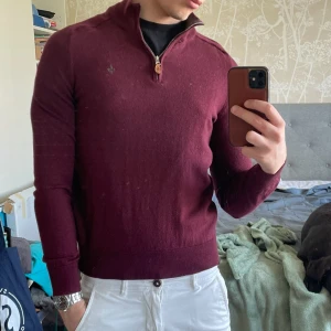 Vinröd merinoullströja med dragkedja från Morris - Vinröd långärmad tröja från Morris i 100% merinoull. Tröjan har halv dragkedja vid halsen och en liten broderad logga på bröstet. Perfekt för lager-på-lager och snygg till både jeans och chinos.