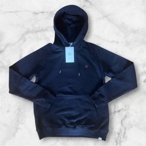 Mörkblå hoodie från Les Deux - Säljer en helt ny med prislapp mörkblå hoodie från Les Deux med broderad orange detalj på bröstet. Tröjan har huva med dragsko och en klassisk känguruficka framtill. Perfekt för en avslappnad look.