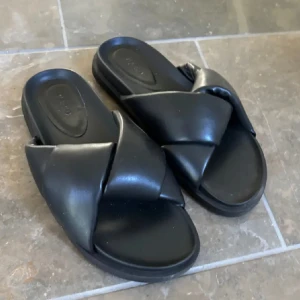 Svarta slip-in sandaler med korsade remmar - Säljer ett par svarta sandaler med breda korsade remmar och platt sula. Från nakd i strl 38. Pris 150 kr❤️Knappt använda 