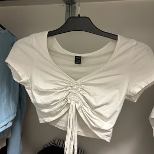 Vit croppad topp med knyt - Säljer en vit croppad topp med snygg knytning framtill och v-ringad hals. Toppen har korta ärmar och är perfekt för varma dagar. Enkel att matcha med både jeans och kjol.