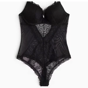 Spetsbody - Säljer en svart bodysuit med spetsdetaljer och meshpaneler. Den har tunna axelband och vadderade kupor för extra stöd. 
