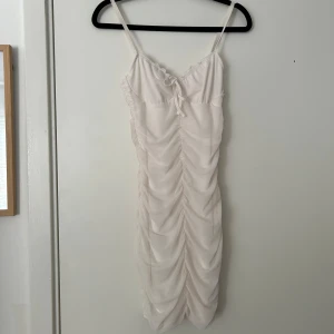 Ljusvit omlottklänning med volang - Säljer en ljusvit kortklänning med smala axelband och omlottdesign. Klänningen har volangdetaljer och rynk längs hela framsidan, samt en liten rosett vid bysten. Perfekt för sommarens alla tillfällen!