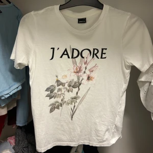 Vit t-shirt med blommigt tryck och text - Vit t-shirt från Ginatricot med trycket 'J'ADORE' och blommotiv framtill. Klassisk rund halsringning och korta ärmar. Perfekt för en avslappnad och trendig look.