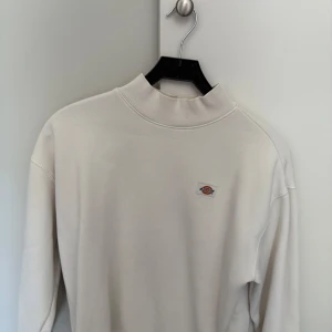 Vit sweatshirt från Dickies - Säljer en stilren vit sweatshirt från Dickies med liten broderad logga på bröstet. Tröjan har hög krage och långa ärmar, perfekt för en enkel och clean look.