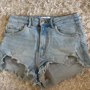Ljusblå jeansshorts från Zara - Säljer ett par ljusblå jeansshorts från Zara med fransiga kanter och råa avslut. Klassisk femficksmodell med knapp och dragkedja framtill. Perfekta för varma dagar och enkel att matcha med olika toppar.