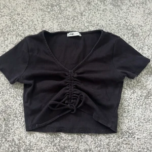 Svart croppad topp med knyt - Svart ribbad croppad topp från New Yorker med v-ringning och knyt framtill. 100% bomull. Kortärmad modell som passar perfekt till jeans eller kjol. Snygg och enkel att styla till olika looks.perfekt för sommaren;)
