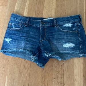 Blå jeansshorts från Abercrombie & Fitch - Säljer ett par blå jeansshorts från Abercrombie & Fitch med slitna detaljer och fransig kant. Klassisk femficksmodell med låg midja och slitningar. Storleken motsvarar ungefär S-M. Pris kan diskuteras! 💕