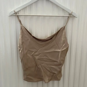 Beige satinlinne - ZARA - Snyggt beige linne i satin med smala axelband och dekorativa knappar i ryggen. Linnet har en lätt glansig yta och en avslappnad passform, perfekt för sommardagar eller att styla lager på lager.