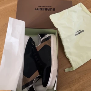 Burberry sneakers med klassiskt rutmönster - Snygga sneakers från Burberry med svart ovandel och klassiskt beige, svart och vitt rutmönster på sidorna. Skorna har vita sulor och svarta snören. Perfekta för dig som vill ha en stilren och lyxig look.