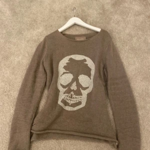 Beige stickad tröja med döskalle från Zadig & Voltaire - Säljer en beige stickad tröja från Zadig & Voltaire med ett stort döskallemotiv i vita stenar framtill. Tröjan har rund halsringning och långa ärmar. Perfekt för dig som gillar edgy detaljer och vill sticka ut. Unik. Säljs inte längre. I nyskick 