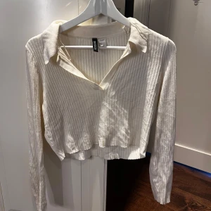 Vit ribbad croppad tröja från Divided - Säljer en vit ribbad croppad tröja från Divided med krage och v-ringning. Tröjan har långa ärmar och är perfekt för lager-på-lager. Enkel och stilren design som passar till mycket.