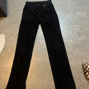 Svarta velourbyxor från Juicy Couture - Mysiga svarta velourbyxor från Juicy Couture med dragsko i midjan och två fickor bak med knappar. Klassisk modell med raka ben och mjukt material, perfekt för en avslappnad stil.
