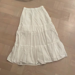 Vit långkjol från Lindex - Säljer en vit långkjol från Lindex med volanger. Kjolen har resår i midjan och är tillverkad i ett lätt och luftigt material, perfekt för varma dagar. Enkel och stilren design som passar till många olika outfits.