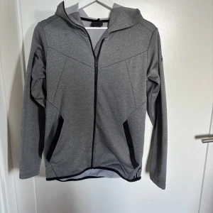 Grå hoodiejacka från Peak Performance - Säljer en grå hoodiejacka från Peak Performance med dragkedja och svarta detaljer vid fickorna. Jackan har lång ärm och huva, perfekt för lager-på-lager och vardagsstil.