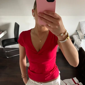 Röd v-ringad topp - Säljer en snygg röd topp med v-ringning och korta ärmar. Toppen har en figurnära passform och är perfekt att matcha med både jeans och kjol. Enkel men stilren design med liten twist vid bysten.
