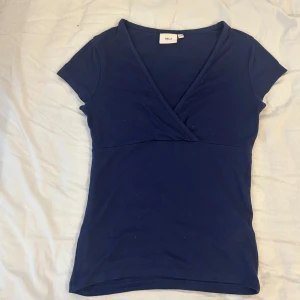 Mörkblå omlottopp från Nelly - Säljer en mörkblå omlottopp från Nelly med korta ärmar och v-ringad hals. Toppen har en figurnära passform och är perfekt för en stilren look.