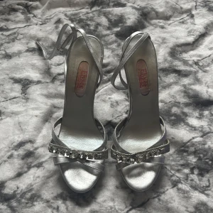 Silverfärgade sandaler med strass från Elysée - Säljer ett par eleganta silverfärgade sandaler från Elysée med smala remmar och glittrande strassdetaljer framtill. Skorna har öppen tå och klack, perfekta för att sticka ut på speciella tillfällen.