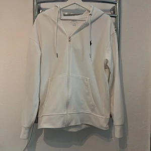 Vit hoodie med dragkedja från Polo Ralph Lauren - Vit hoodie från Polo Ralph Lauren med dragkedja framtill och klassisk logga broderad på bröstet. Tröjan har huva med dragsko och en stor magficka. Perfekt för en avslappnad stil. Hoodien är i toppskick 10av10!!