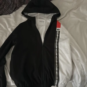 Svartvit sportig vindjacka från Lacoste Sport - Säljer en snygg svartvit vindjacka från Lacoste Sport med huva och dragkedja. Jackan har en röd detalj på axeln, Lacoste-logga på bröstet och sportiga band längs dragkedjan. Perfekt för en sportig stil.