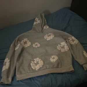 Denim tears hoodie - Säljer en grå Denim tears hoodie med vita blommor tryckta över hela tröjan. Tröjan har huva och långa ärmar, samt en klassisk magficka framtill. Perfekt för dig som gillar en avslappnad och trendig stil.