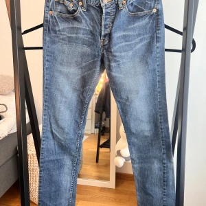 Levis 501 blå jeans strl W30 L32 - Ett par blå jeans med fickor fram och bak. Jeansen har en knappgylf och en brun läderpatch bak i midjan.   Ett par klassiska Levi's jeans med en tidlös design. Jeansen har en traditionell femficksmodell och en knappgylf. Denimen har en snygg tvätt som ger en avslappnad och använd look. Dem är lågmidjade och i en straight modell.