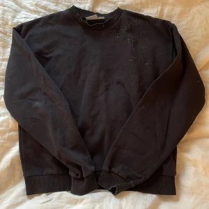 Svart sweatshirt med stjärndetaljer från Gina Tricot - Svart sweatshirt från Gina Tricot med rund halsringning och ribbade muddar. Framsidan är dekorerad med svarta stjärnor i små glittriga pärlor. Perfekt för dig som gillar en enkel men ändå unik look.