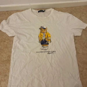 Vit t-shirt med Polo Bear från Ralph Lauren - Vit t-shirt från Ralph Lauren med ikoniska Polo Bear-trycket på bröstet. Björnen har gul jacka, keps och sneakers. Klassisk rund hals och korta ärmar. Perfekt för dig som gillar streetwear och snygga prints.