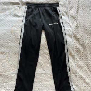 Svarta träningsbyxor från Palm Angels med vita revärer - Palm Angels svarta träningsbyxor med vita revärer längs sidorna och logga tryckt på vänster lår. Byxorna har elastisk midja och raka ben, perfekt för en sportig och trendig look.