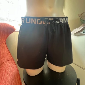 Svarta träningsshorts från Under Armour - Svarta träningsshorts från Under Armour med bred resår i midjan och logga runtom. Modellen är loose fit och har snörning i midjan. Perfekta för träning och sportiga aktiviteter.