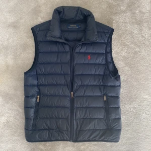 Ralph Lauren väst - Snygg mörkblå dunväst från Polo Ralph Lauren med dragkedja och två fickor framtill. Västen har en klassisk ståkrage och den ikoniska röda loggan broderad på bröstet. Perfekt för lager-på-lager-stil.