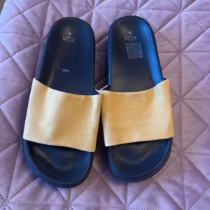 Gula sandaler från VOX storlek 40 - Säljer ett par snygga sandaler från VOX i storlek 40. De har en bred gul rem över foten och svart sula med platt design. Perfekta för varma dagar och enkel att matcha med olika outfits.