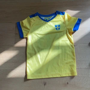 Svensk fotbollströja med nummer 10 - Gul fotbollströja med blå detaljer och Sveriges landslagsemblem på bröstet. På ryggen står det 'Sverige' och siffran 10 i blått. Tröjan har korta ärmar och är tillverkad i ett lätt, ventilerande material. Perfekt för fotbollsfans!