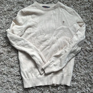 Beige stickad tröja från Morris - Säljer en stilren beige stickad tröja från Morris. Tröjan har rund halsringning och diskret logga broderad på bröstet. Perfekt för en klassisk och enkel look. Långa ärmar och ribbade muddar vid ärmslut och nederkant.