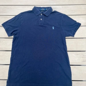 Blå piké från Polo Ralph Lauren - Säljer en klassisk blå pikétröja från Polo Ralph Lauren med broderad logga på bröstet. Tröjan har korta ärmar, krage och knappar framtill. Modellen är custom slim fit och passar perfekt till en avslappnad stil. Den är lite använd men superfint skick!!