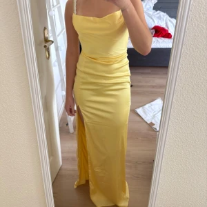 Gul långklänning med slits och smala axelband - Säljer en lång gul klänning med smala axelband och draperad urringning. Klänningen har en hög slits framtill och är tillverkad i ett glansigt, satinliknande material. Perfekt för dig som vill sticka ut med en färgglad look. Är även helt ny!