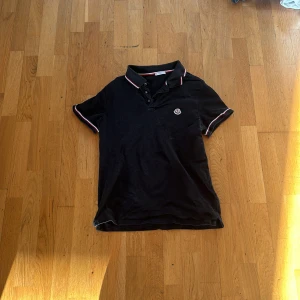 Svart pikétröja från Moncler - Säljer en svart pikétröja från Moncler med klassisk krage och knappar framtill. Tröjan har Moncler-logga på bröstet och rödvita detaljer på krage och ärmslut. Perfekt för en stilren och sportig look.