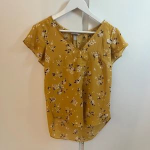 Gul blommig blus från H&M - Söt gul blus från H&M med vitt och svart blommönster. Blusen har korta ärmar, v-ringning och är tillverkad i ett lätt och luftigt material. Perfekt för varmare dagar och ger en fräsch look.
