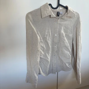 Beige linneskjorta från H&M - Snygg beige skjorta i linne från H&M med klassisk krage och bröstficka. Skjortan har långa ärmar och knäppning framtill samt indragen midja Perfekt för en stilren och avslappnad look. Helt oanvänd, endast testad