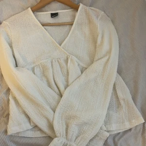Vit omlottopp från Gina Tricot - Säljer en vit omlottopp från Gina Tricot i strukturerat tyg. Toppen har lång ärm med resår vid ärmslut och v-ringad omlottringning. Perfekt för dig som gillar en luftig och stilren look. Säljer eftersom den bara hänger i garderoben och ej kommer till användning ❤️