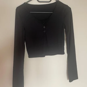 Svart croppad långärmad topp med knappar - Säljer en svart croppad långärmad topp med v-ringning och tre små knappar framtill. Perfekt att matcha med höga jeans eller kjol för en stilren look.🖤