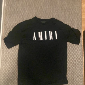 Svart t-shirt från Amiri - Säljer en svart t-shirt från Amiri med stor vit logga tryckt över bröstet. Over size passform och rund halsringning. Perfekt för dig som gillar stilrena och trendiga plagg med tydlig branding.